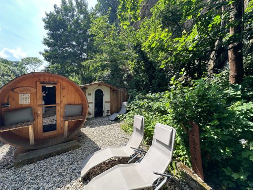 Holzfass-Sauna in einem grünen Garten, umgeben von Bäumen und zwei Liegen.